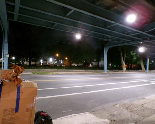 The image shows a dimly lit urban street beneath an elevated structure, with cardboard boxes and a trash bag visible in the foreground.