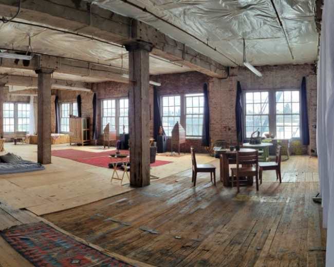 The image shows a spacious, industrial-style room with wooden floors, large windows, and scattered furniture, including chairs and tables, in a creative workspace.