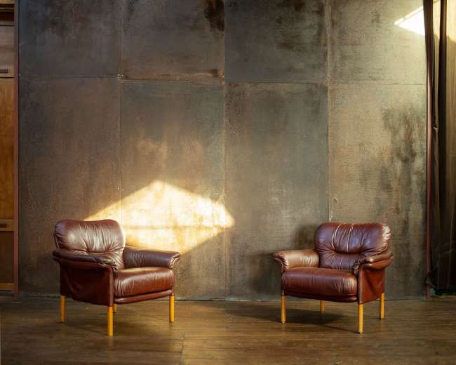 Two brown leather chairs are positioned facing each other against a textured metal wall, with sunlight illuminating one chair.