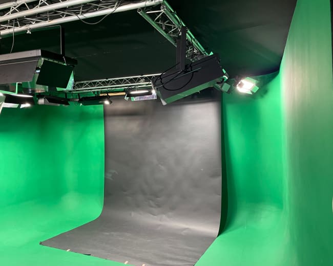 The image shows a green screen setup in a studio, equipped with lighting fixtures and a black backdrop.