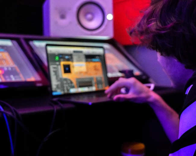 A person is working on a laptop surrounded by audio equipment in a music production studio.