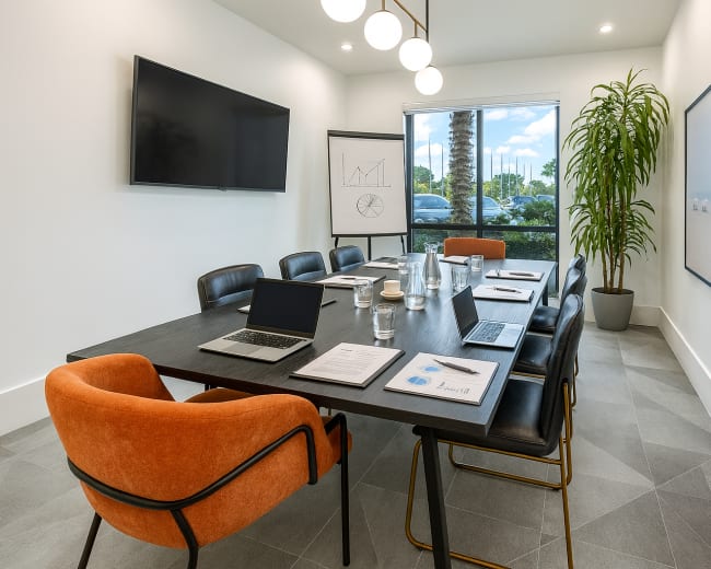 The image shows a modern conference room equipped with a large table, multiple laptops, a presentation board, and a view of palm trees outside.