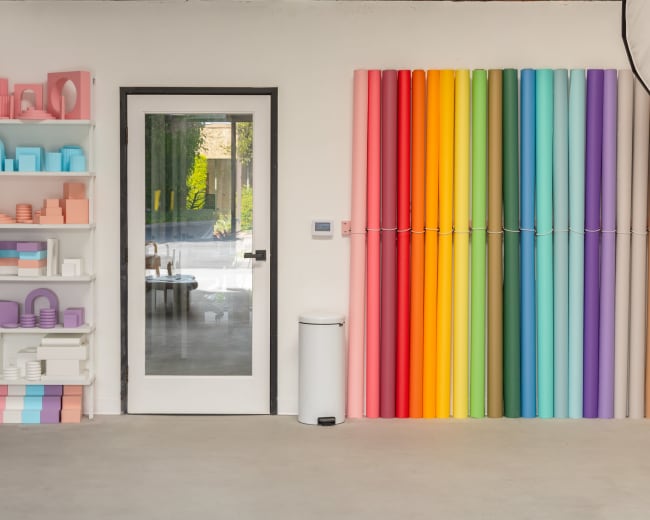 The image shows a modern interior with a white wall featuring colorful rolled paper and a shelf displaying various geometric shapes in pastel colors, adjacent to a glass door.