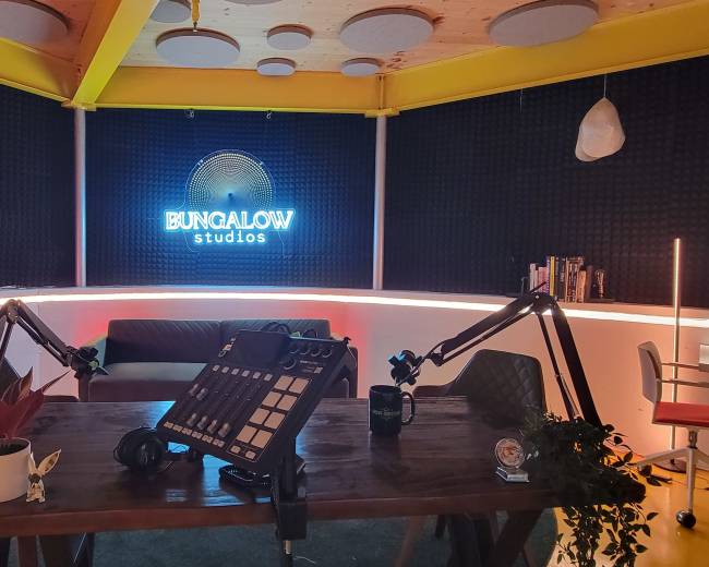 The image shows a recording studio with a neon sign that reads "Bungalow Studios," featuring a central table, microphone stands, and seating arrangements against a backdrop of soundproofing panels.