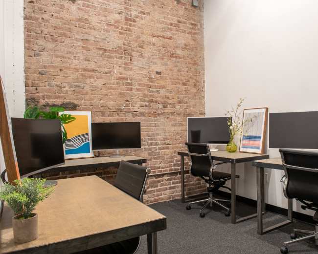 The image shows a modern office workspace with four black computer monitors arranged at two desks against a brick wall.