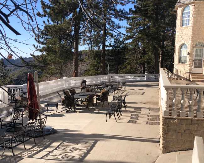 A stone patio with several tables and chairs sits beside a building, surrounded by tall trees and clear blue skies.