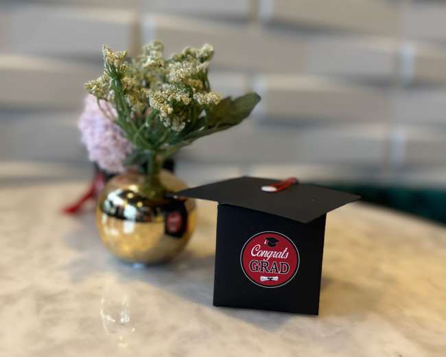 A small graduation cap decoration with a "Congrats Grad" sticker sits next to a floral arrangement on a marble table.