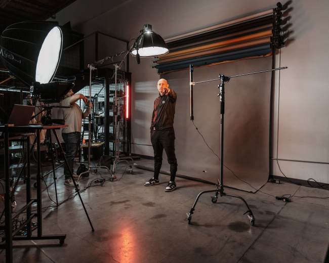 A person stands in a photography studio, pointing towards the camera while surrounded by lighting equipment and a backdrop.