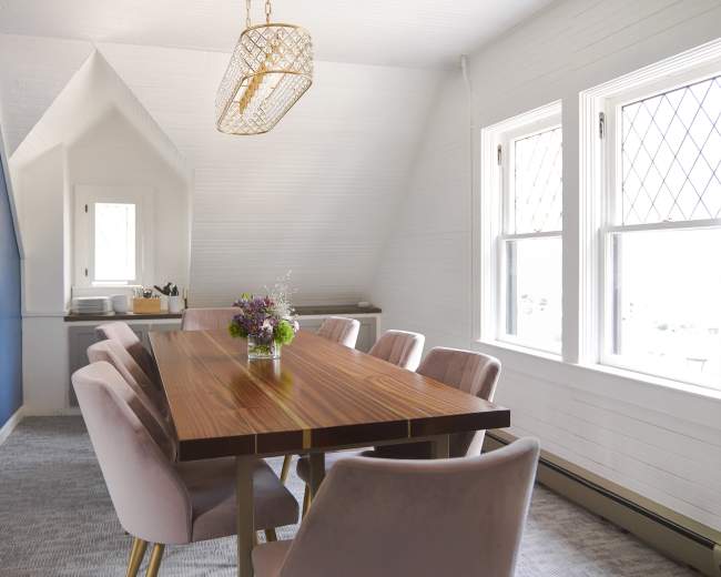 A modern dining room features a long wooden table surrounded by pink upholstered chairs, illuminated by a chandelier, with large windows letting in natural light.