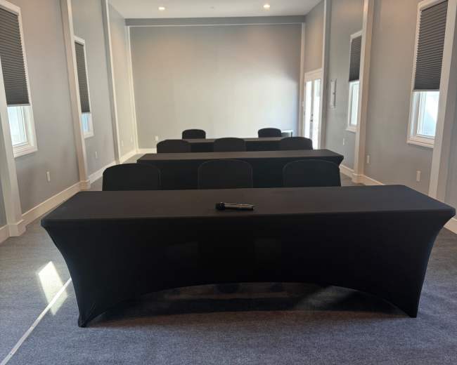 The image shows a conference room with a long table covered in a black tablecloth, surrounded by several chairs, and illuminated by natural light coming through windows.