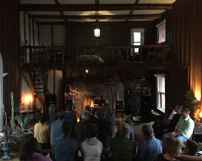 A group of people sit in a cozy room with a stone fireplace, watching a performer play the violin while another musician sits nearby.