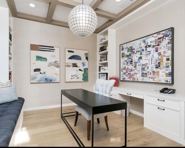 The image shows a modern home office with a central black table, a blue sofa along one wall, two abstract paintings, and a bulletin board filled with colorful papers and notes.