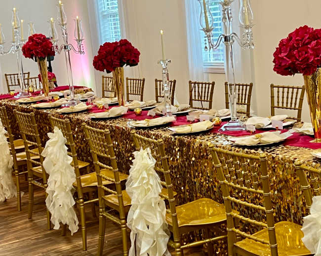 A long dining table is set with gold and white decor, featuring red floral centerpieces and elegant tableware.