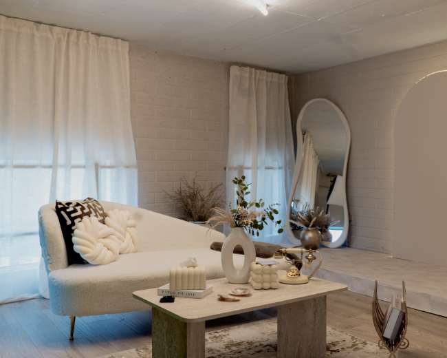 The image depicts a modern living room with a light-colored sectional sofa, a minimalistic coffee table, and decorative elements against a backdrop of soft curtains and textured walls.