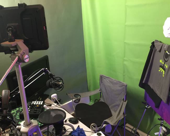 A small studio setup featuring a green screen, a microphone on a boom arm, a computer monitor, a folding chair, and clothing hanging on a rack.