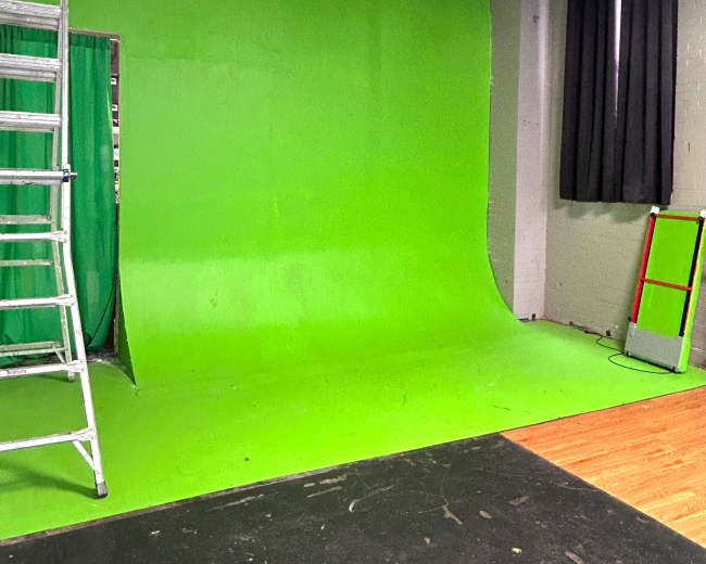 The image shows a green screen setup in a studio, accompanied by a ladder and large black curtains.