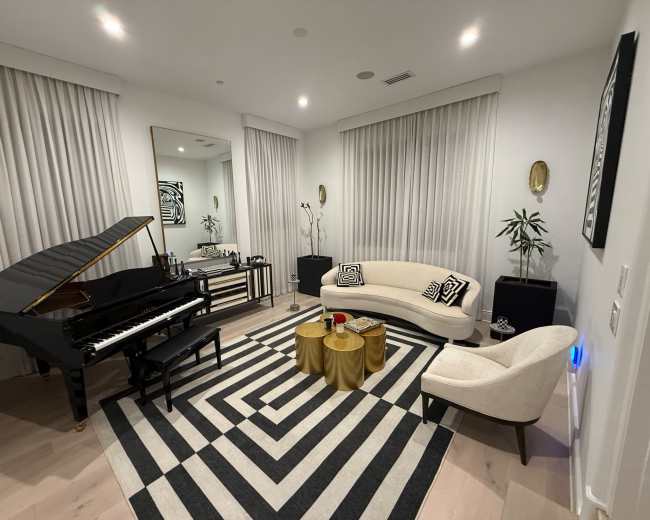 The image shows a modern living room featuring a grand piano, a curved sofa, two gold coffee tables, and large windows covered with sheer curtains.