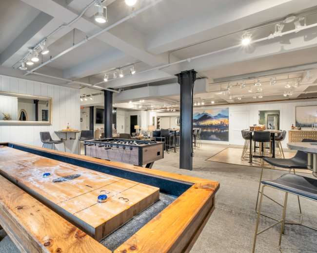 The image shows a modern recreational room featuring a shuffleboard table and a foosball table, with a bar area and art on the walls.
