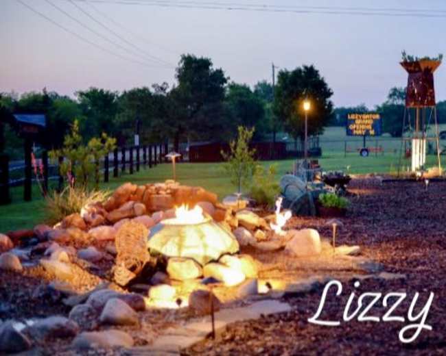 A couple walks along a stone pathway illuminated by fire features in a landscaped area with trees and a sign in the background.