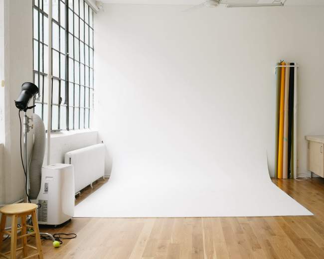 The image shows a bright, minimalistic studio space with a white backdrop, wooden flooring, and a stool.