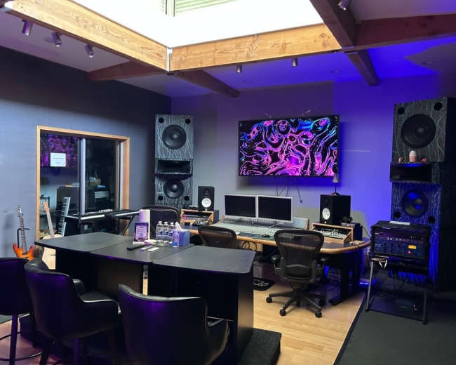 The image shows a modern music studio featuring a mixing desk, computers, large speakers, and various musical instruments along the walls.