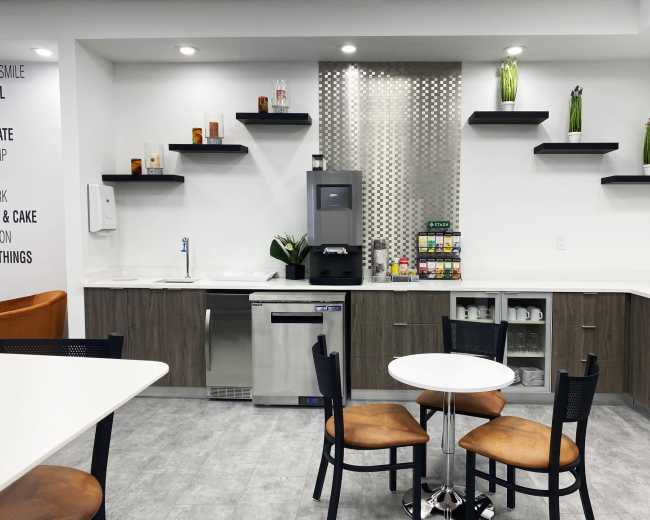 The image shows a modern kitchenette with a countertop, coffee machine, shelves displaying decorative items, and a small round table with four chairs.