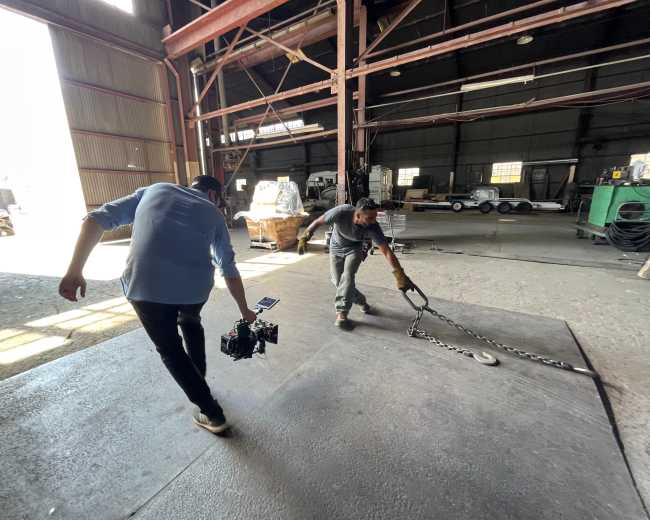 A man pulls a large metal chain on a workshop floor while another man carries a camera on a gimbal.