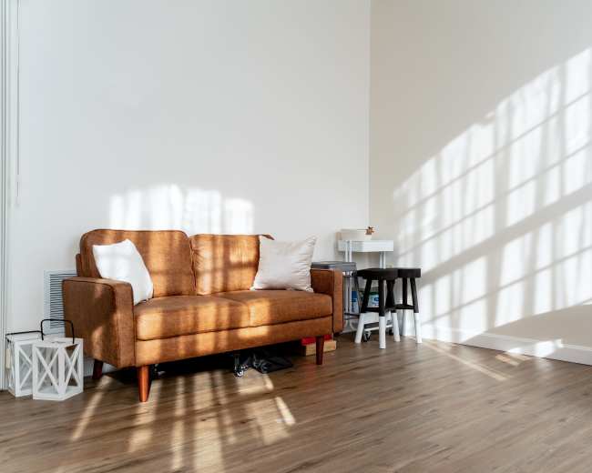 A brown sofa with a white pillow sits against a wall, casting shadows from nearby window panes on the floor.
