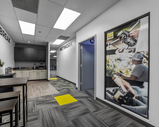 A modern office hallway features a kitchen area on one side and a large poster of a photographer on the other, with yellow and gray floor tiles.