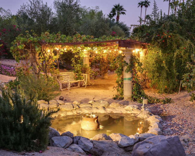 A stone pond with a rustic pot sits at the center of a garden featuring a vine-covered pergola illuminated by hanging lights.