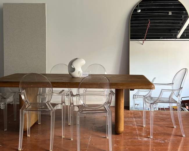 A wooden dining table is surrounded by clear acrylic chairs in a minimalist room with white walls and a large mirror.
