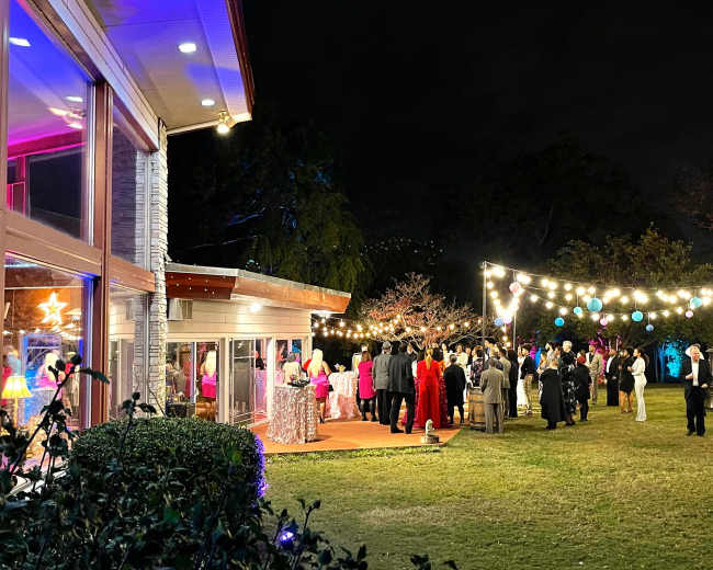A gathering of people is taking place outdoors at night, with colorful lights hanging above and a house visible in the background.