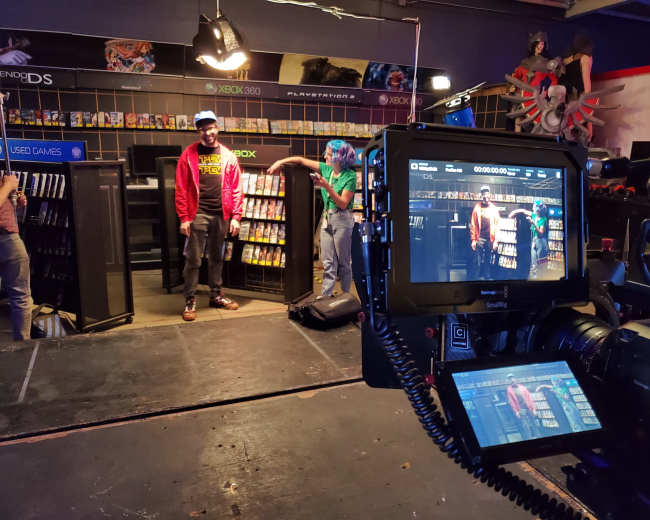 A film set featuring two people standing in a video game store aisle, with shelves filled with video games and a camera focused on them.