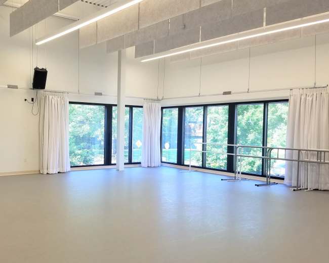 The image shows a large dance studio with white walls, smooth flooring, and large windows letting in natural light.