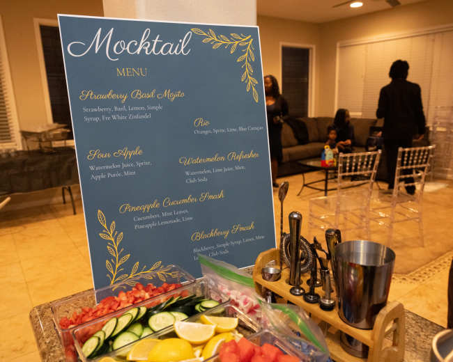 A mocktail menu displayed on a blue board lists various drink options alongside a preparation station featuring fresh fruits and garnishes.