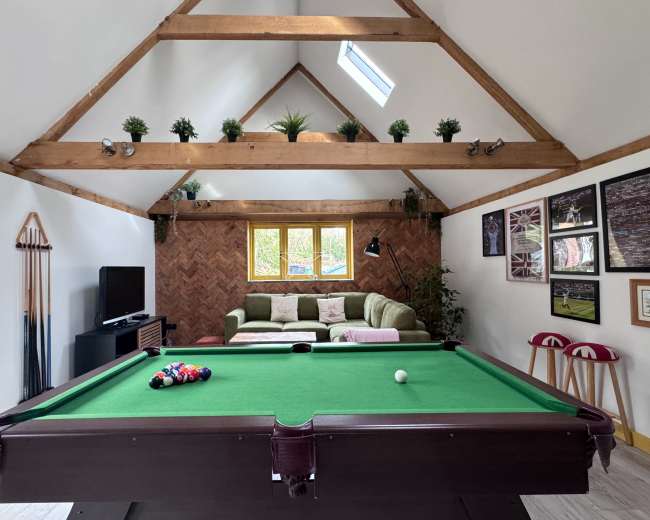 The image shows a games room with a green pool table in the foreground, a brown herringbone wall, and a seating area with a green sofa and plants against a white wall.