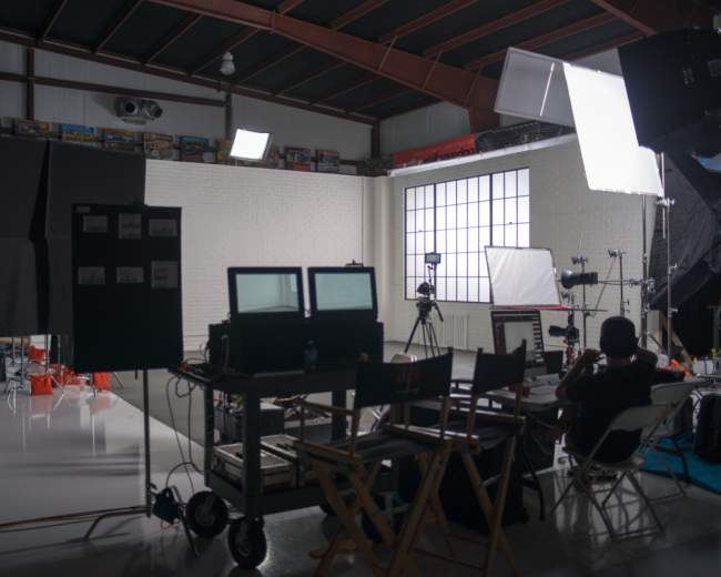 A film set features multiple cameras, lighting equipment, and two monitors arranged on a cart in a spacious studio with a large window.