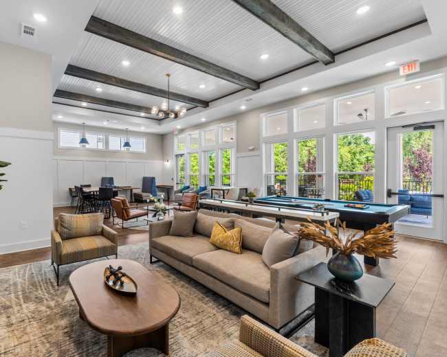 The image shows a spacious community lounge area featuring modern furniture, large windows, and an open layout with recreational tables.