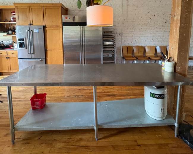 A stainless steel worktable is centrally located in a large kitchen, with wooden cabinets and shelves in the background, and a pendant light hanging above it.