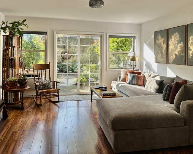 The image shows a bright living room with a sectional couch, a rocking chair, and large windows displaying a garden view.