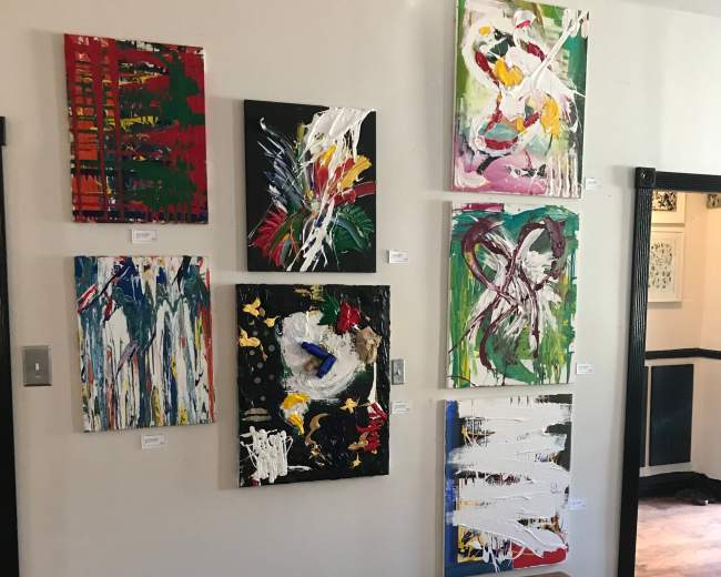 A wooden bench sits in front of a wall featuring eight abstract paintings in various colors and styles.