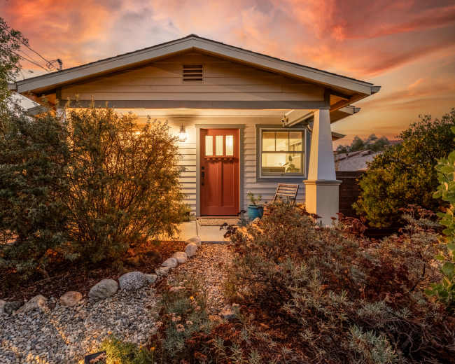 A small house with a brown front door is surrounded by landscaped gardens and rocks under a colorful sunset sky.