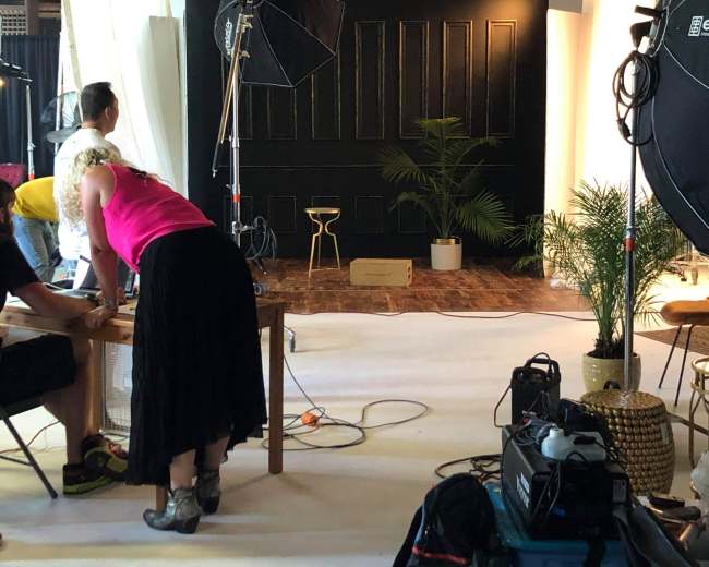 A studio setup features a dark wall with a stool, surrounded by lighting equipment and two people preparing for a shoot.