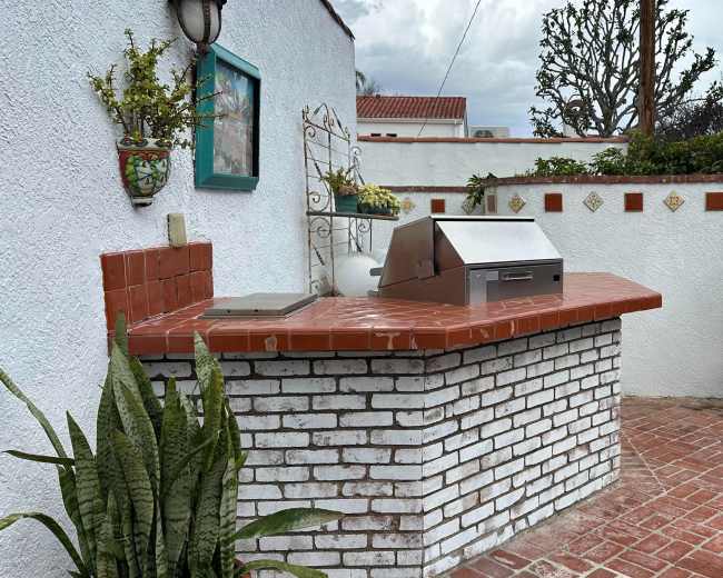 The image shows an outdoor area featuring a brick grill station with a stainless steel grill, surrounded by planters and a white wall adorned with decorative tiles.