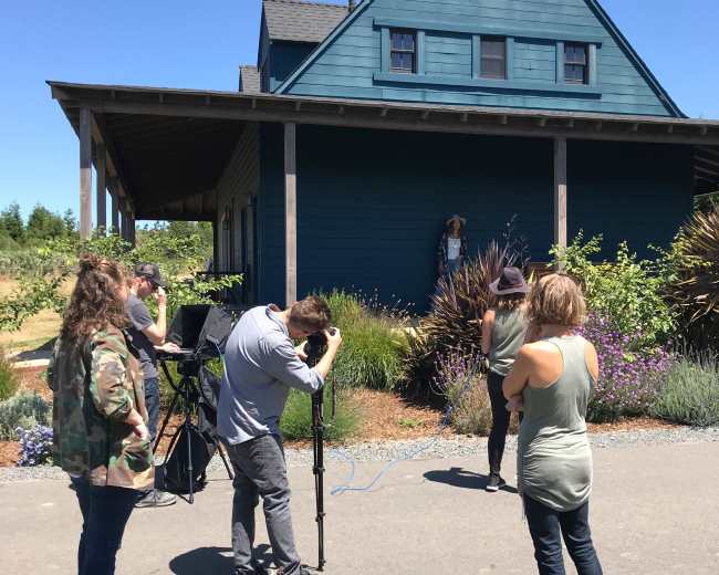 A group of people are staging a photoshoot in front of a blue house surrounded by greenery and flowers.