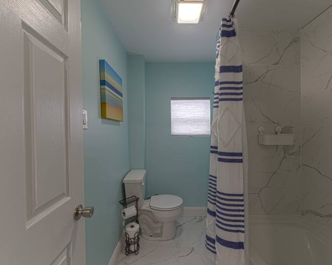 A bathroom features a toilet, a bathtub with a shower curtain, a small window, and a colorful painting on the wall.