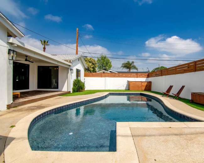The image shows a swimming pool surrounded by a patio, with two lounge chairs and a wooden storage box beside a white fence.
