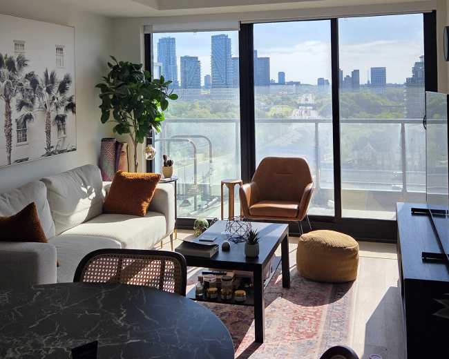 The image shows a modern living room with a view of city skyscrapers through large glass doors, featuring a cozy seating area and a round dining table.