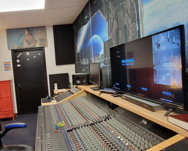 A recording studio with a large mixing console, a computer monitor displaying audio software, and a blue leather chair in a room featuring soundproofing panels on the walls.