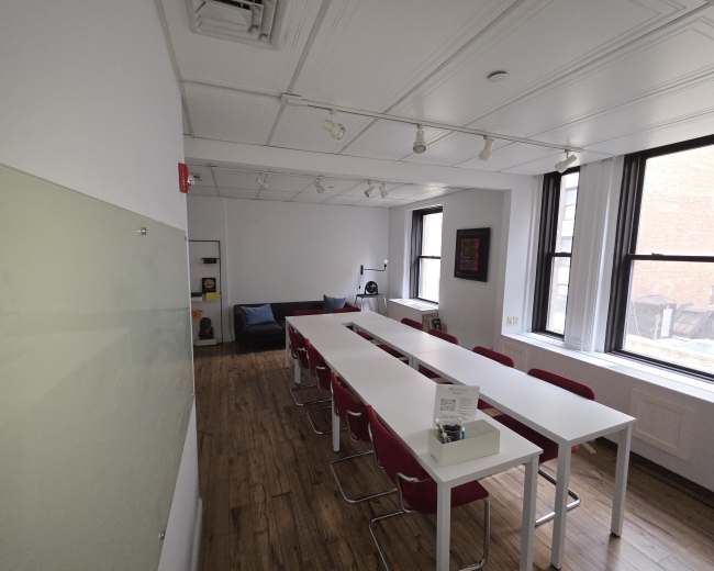 The image shows a bright, modern office space with a long white table surrounded by red chairs and a couch in the corner.